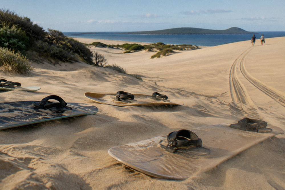 Sandboarding boards resting on a smooth sand slope at the base of a tall dune, with vehicle tracks running across the sand