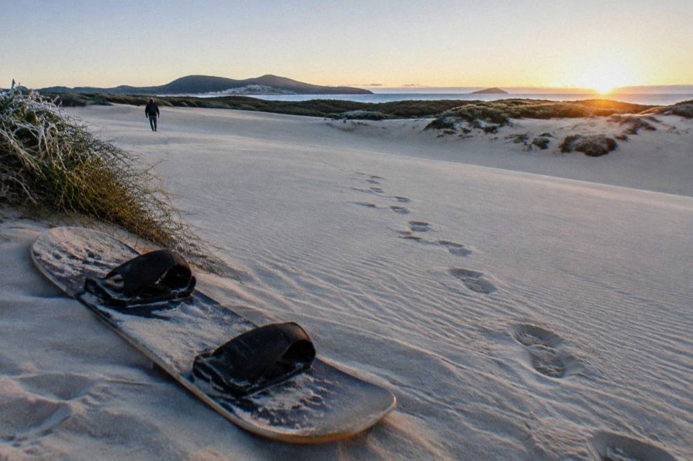 A sandboard resting on a smooth dune slope with footprints trailing down the sand