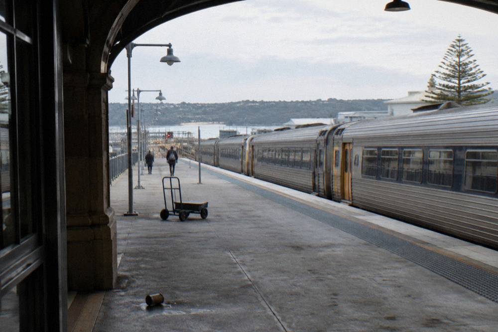 A country rail platform beside open ground with a train waiting under a wide sky