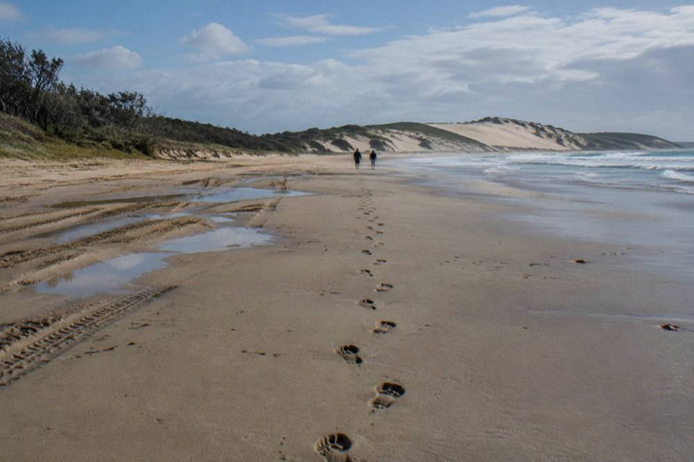 A line of footprints crossing a broad slope of pale sand toward distant dune ridges