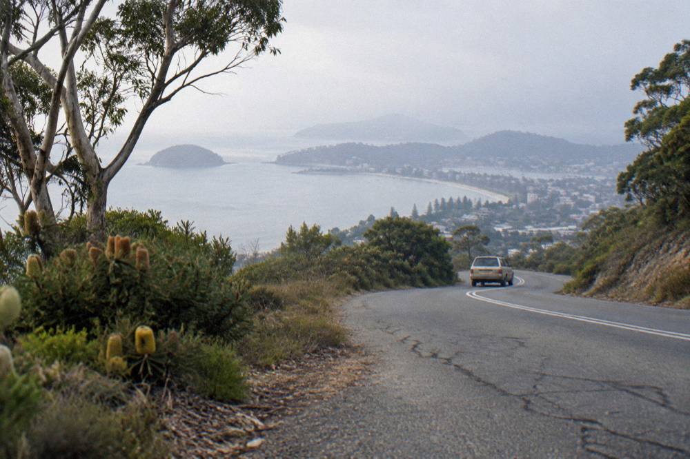 A coastal road curving past bushland toward a distant bay town