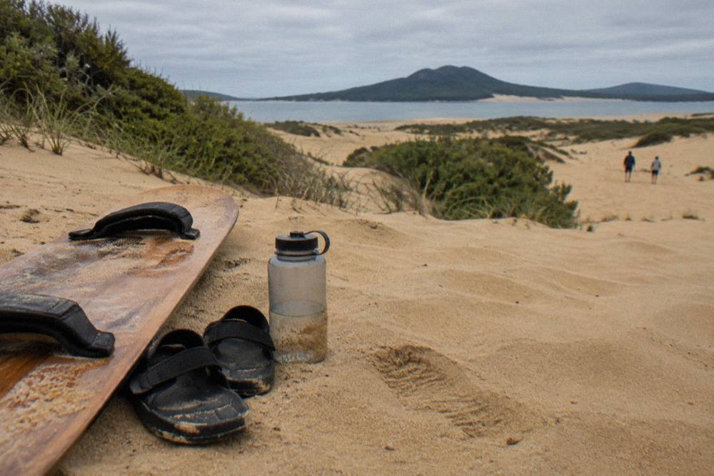 A pair of sandals and a water bottle resting beside a sandboard on golden dune sand