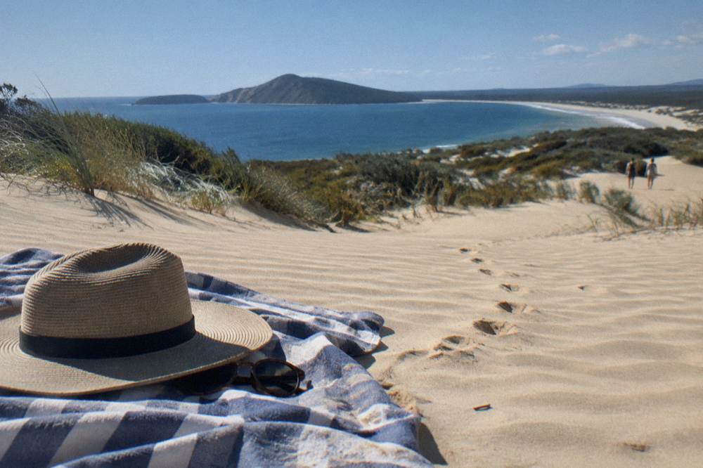 A wide brim hat and sunglasses resting on a towel on golden dune sand