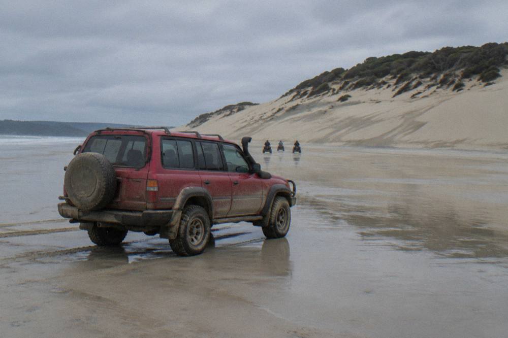 A red 4WD tour vehicle parked on wet sand at the base of the Stockton Bight dunes under a grey sky