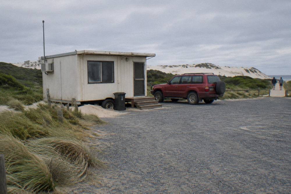 A gravel meeting area with a small reception trailer and a red four wheel drive vehicle waiting beside open dunes