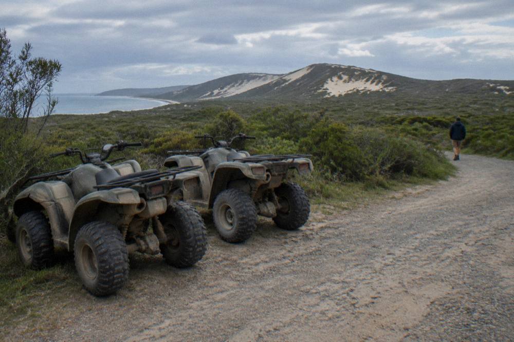 Two quad bikes lined up at the edge of a sandy track with dune ridges in the distance