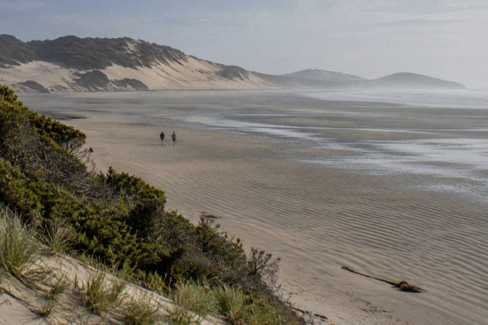 A broad flat beach at low tide with sand dunes rising behind it and a light haze over the water
