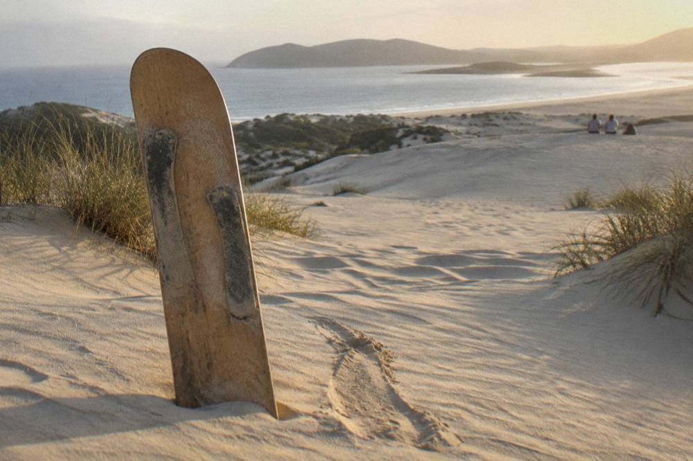 A wooden sandboard resting on the crest of a smooth sand slope with dune ridges beyond