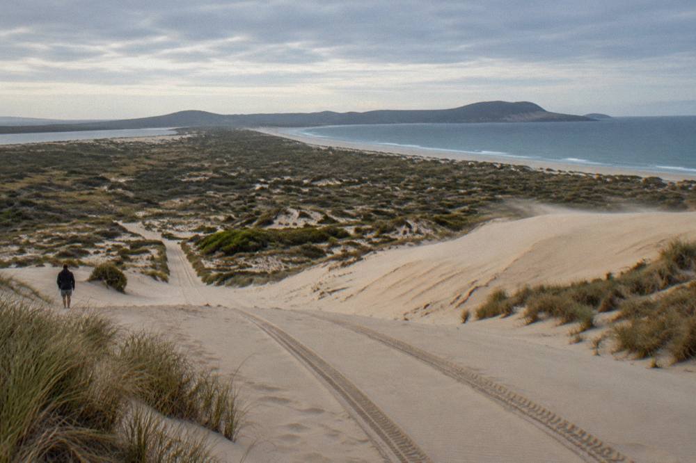 A line of tyre tracks crossing a broad slope of pale sand dunes under open sky