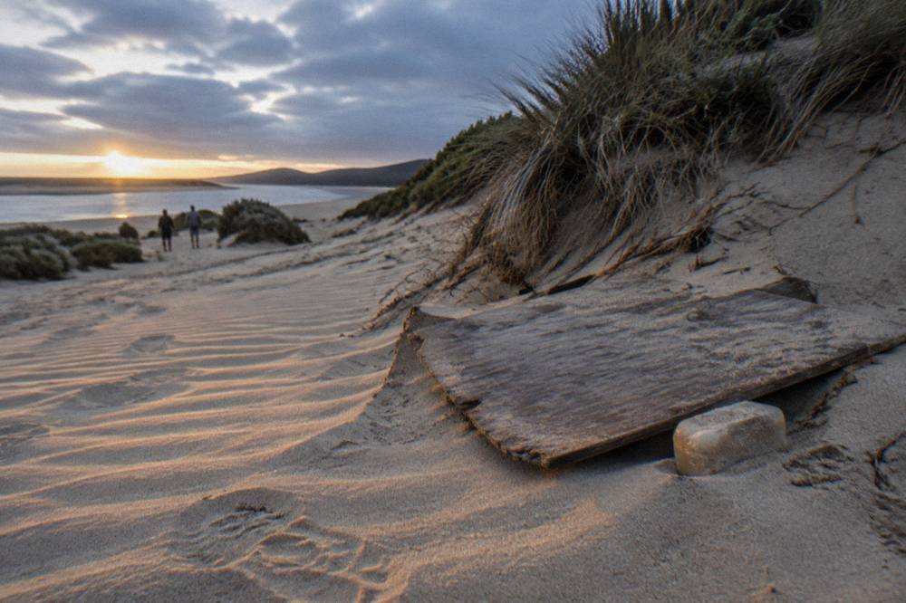 A weathered wooden board resting on rippled sand at the base of a tall dune