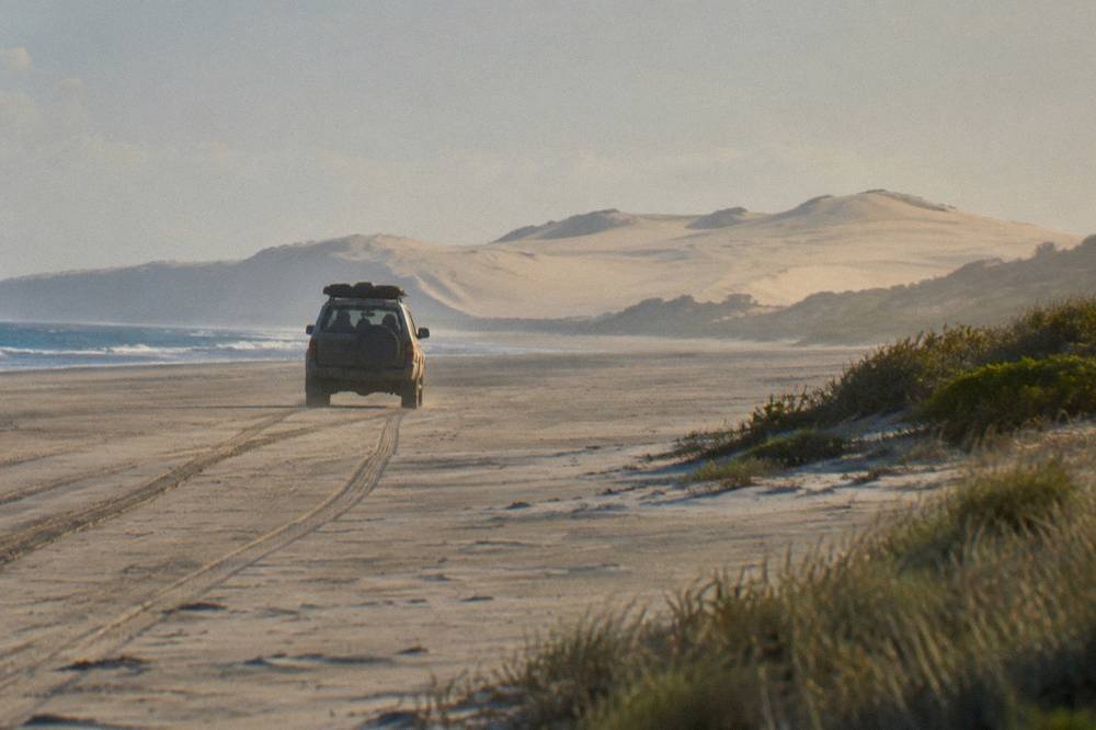 A four-wheel-drive vehicle with passengers crossing a wide flat stretch of sand toward distant dunes